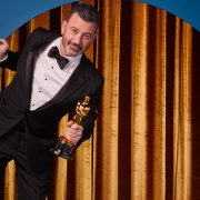 Jimmy Kimmel wears a tuxedo and a bowtie and holds an Oscar in his left hand. He is standing in front of gold curtains and is leaning out of a circular cutout in a blue wall.