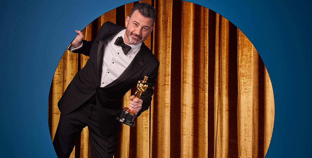 Jimmy Kimmel wears a tuxedo and a bowtie and holds an Oscar in his left hand. He is standing in front of gold curtains and is leaning out of a circular cutout in a blue wall.