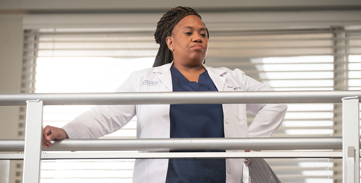 Dr. Miranda Bailey, played by Chandra Wilson, wears a white lab coat and blue scrubs. She has a stack of papers in her left hand and rests her right hand on a silver railing.