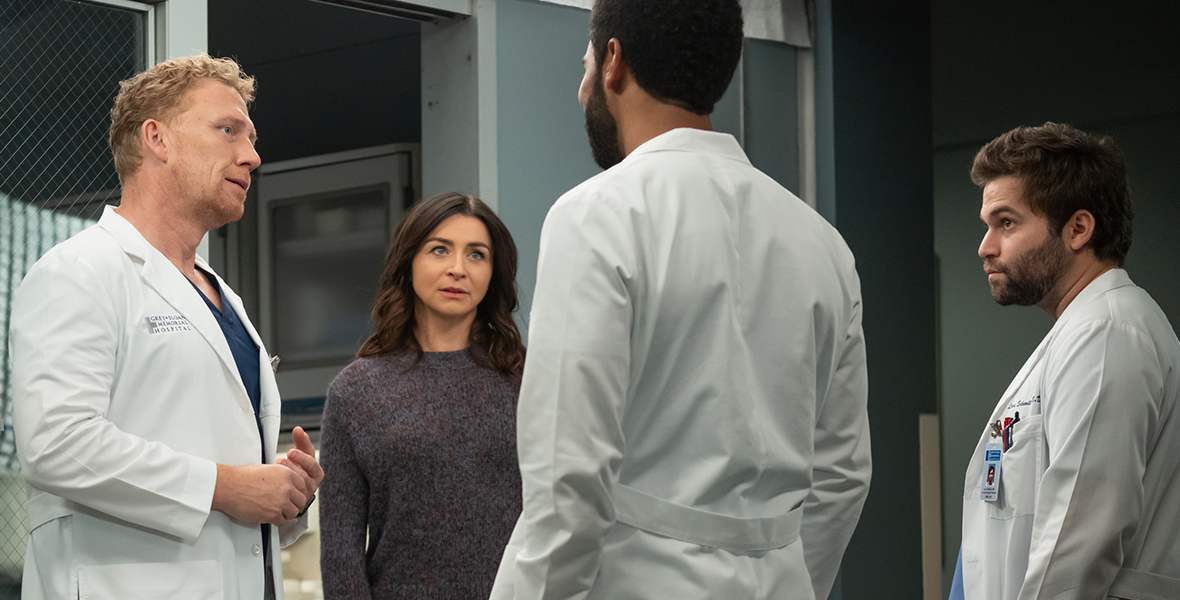 Dr. Owen Hunt, played by Kevin McKidd, Dr. Amelia Shepherd, played by Caterina Scorsone, Dr. Winston Ndugu, played by Anthony Hill, and Dr. Levi Schmitt, played by Jake Borelli, stand in a circle outside of a hospital room. The male doctors are wearing scrubs and white lab coats, while Dr. Shephard is wearing a purple wool sweater and black pants.