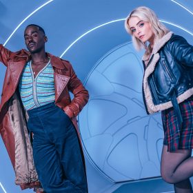 Ncuti Gatwa (left) and Millie Gibson (right) are standing inside a life-size blue cylinder illuminated by neon white light streaks. Gatwa wears a burgundy leather trench coat, gray sneakers with white accents, blue velvet pants, and a zipped-up knit top with colorful stripes. Gibson sports black combat boots, black sheer tights, a checkered mini skirt, and a shearling black leather jacket.