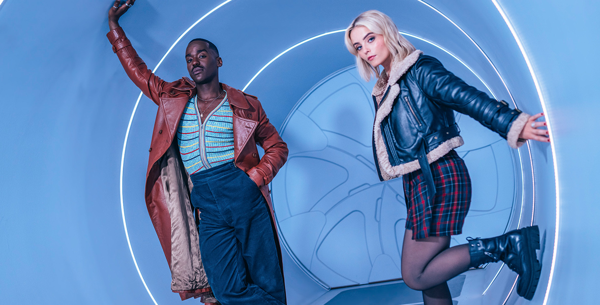 Ncuti Gatwa (left) and Millie Gibson (right) are standing inside a life-size blue cylinder illuminated by neon white light streaks. Gatwa wears a burgundy leather trench coat, gray sneakers with white accents, blue velvet pants, and a zipped-up knit top with colorful stripes. Gibson sports black combat boots, black sheer tights, a checkered mini skirt, and a shearling black leather jacket.