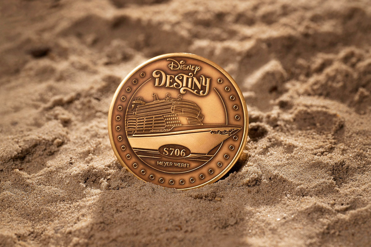 A gold Disney Destiny Keel coin sits in the sand.