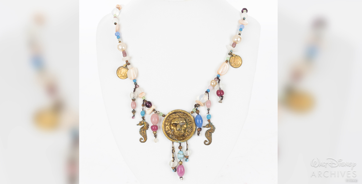 The necklace worn by Madison (Daryl Hannah), pictured around her neck in the image at the top of this story, is seen here displayed against a white background. The beaded necklace, now in the care of the Walt Disney Archives. is adorned with seahorse charms, and ancient coins, along with a bronze-colored medallion depicting a lion’s head.
