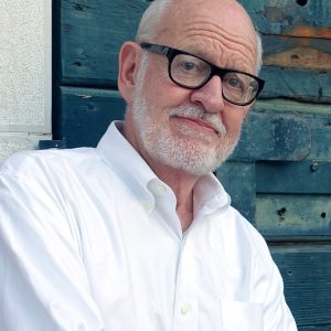 Disney Legend Frank Oz sports a white beard and glasses, wearing a white button-down shirt, standing with his arms crossed against a backdrop of a brick wall and a weathered wooden door.