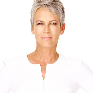 Disney Legend Jamie Lee Curtis sports short silver hair, wearing subtle makeup and small stud earrings, looking directly at the camera with a neutral expression against a plain white background.