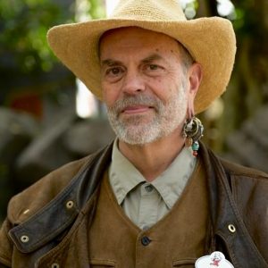 The image features Disney Legend Joe Rohde. He is seen wearing a straw hat and has a distinguished beard. Joe Rohde is also notable for his unique earring, which includes multiple charms and beads.