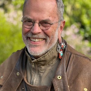 The image features Disney Legend Joe Rohde. He is seen wearing a straw hat and has a distinguished beard. Joe Rohde is also notable for his unique earring, which includes multiple charms and beads.