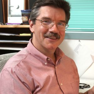 Disney Legend Mark Henn sports glasses and a mustache, wearing a light pink shirt, smiling at the camera while seated at a desk. The background includes a shelf with various items and a window with blinds.