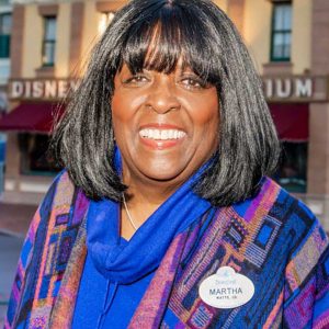 Disney Legend Martha Blanding sports shoulder-length dark hair, wearing a blue and patterned shawl with a Disneyland name tag, smiles while standing in front of a background of shops and buildings.