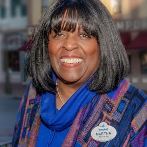 Disney Legend Martha Blanding sports shoulder-length dark hair, wearing a blue and patterned shawl with a Disneyland name tag, smiles while standing in front of a background of shops and buildings.