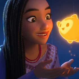 In a scene from Wish, Asha (left) stands in awe, gazing at the mesmerizing Star (right). Asha is dressed in a stunning purple ensemble—a long-sleeved lavender dress adorned with a two-tone purple triangle-inspired design, accentuated by magenta threaded seams and intricate golden decorations adorning the V-cut neckline. The backdrop depicts a serene nighttime setting, with the sea visible in the background and fireworks descending into the ocean.
