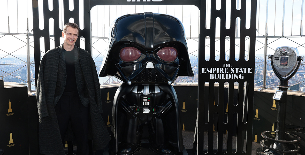 Hayden Christensen sits beside a large sculpture of Darth Vader inside the Empire State Building. He wears a black sweater, pants, loafers, a dark gray blazer, and coat. Above the sculpture is a black arch adorned with "Star Wars," "THE EMPIRE STATE BUILDING," and a Lucasfilm trademark, against the backdrop of the New York City skyline.