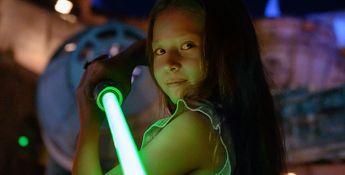 In a poster for the Star Wars Season of the Force event, a young girl is holding a green lightsaber. From top to bottom, the poster reads: “Star Wars Season of the Force,” “4.5.24-6.2.24.”