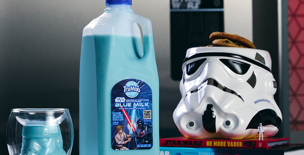 In a kitchen, a TruMoo Star Wars Blue Milk product (left) is on a table. To its right is a Stormtrooper Cookie Jar holds two cookies, resting on two books. 