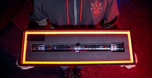 A boy wearing a black Darth Vader hoodie with red detailing is holding up the Darth Maul Legacy LIGHTSABER Set inside a box with neon yellow details.