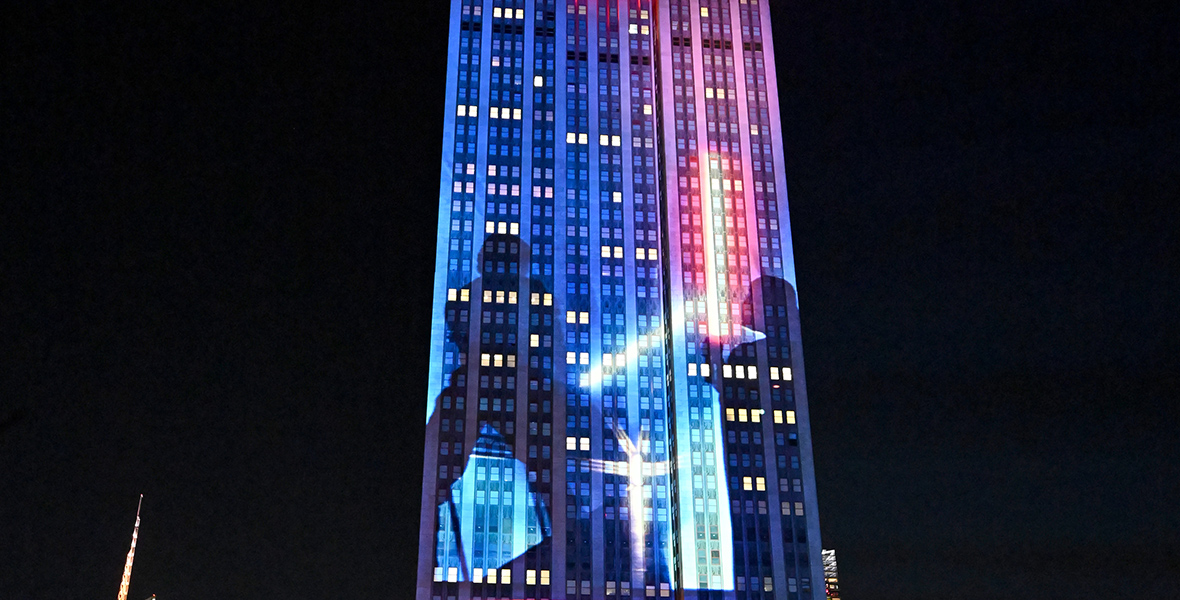 The Empire State Building illuminated, depicting two Star Wars characters engaged in combat with blue and red lightsabers. The building is adorned with hues of red, blue, and purple. The nighttime skyline of New York City forms the background of the photo.
