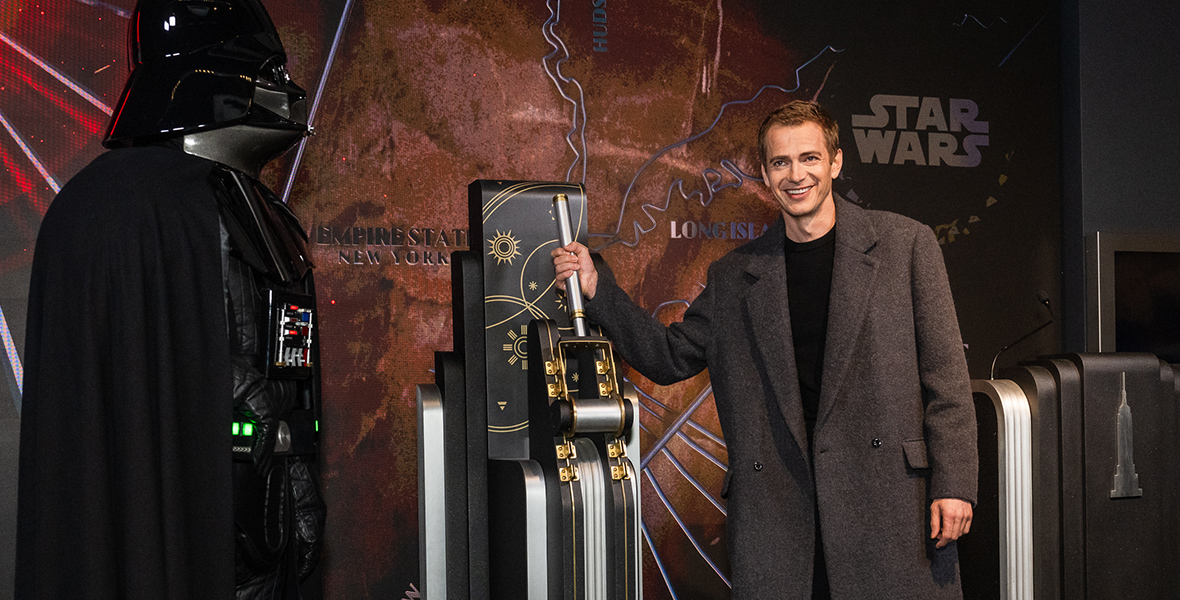 A life-size doll of Darth Vader (left) and Hayden Christensen (right) is about to pull a lever that lights up the Empire State Building. Christensen is wearing a black crewneck sweater and a dark gray coat. 
