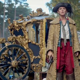 In a scene from the series Renegade Nell, Nell (Louisa Harland) is standing in front of a gold carriage in the middle of the woods. She’s wearing a pirate-style black hat, a white shirt, red pants, brown pirate boots, and a mustard-colored coat. She holds a pistol in her right hand.