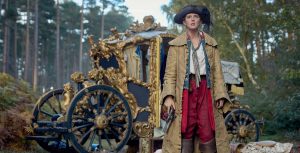 In a scene from the series Renegade Nell, Nell (Louisa Harland) is standing in front of a gold carriage in the middle of the woods. She’s wearing a pirate-style black hat, a white shirt, red pants, brown pirate boots, and a mustard-colored coat. She holds a pistol in her right hand.