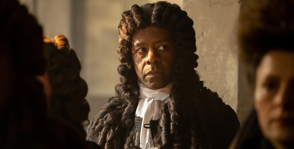 In a scene from the series Renegade Nell, Robert Hennessey, the Earl of Poynton (Adrian Lester) is wearing a brown powdered wig and looking to his left. He’s also wearing a black steampunk-style jacket with gold details and a white point collar dress shirt.