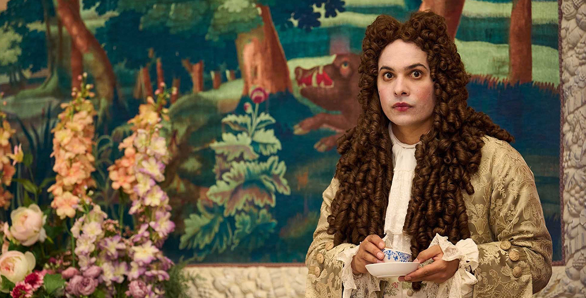 In a scene from the series Renegade Nell, Charles Devereux (Frank Dillane) is wearing a brown powdered wig, a gold steampunk-style jacket, and a white point collar dress shirt. He is holding a white teacup and plate. In the background is a vibrant painting of a forest and a boar, and to Devereux’s right are flowers.