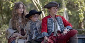 In a scene from the series Renegade Nell, Roxy Jackson (Bo Bragason), George Jackson (Florence Keen), and Nell Jackson (Louisa Harland) are sitting on a carriage. Roxy is wearing a checkered dress with floral patterns and a beige coat with fur trims; George is wearing a black hat, round glasses, a looser-cut coat with patterns, an open-necked white shirt, brown pants that reach her knees, and beige socks; Nell is wearing a pirate-style black hat, a white shirt, red pants, brown pirate boots, a brown belt, and a red and blue Regency tailcoat. She is also holding the reins of the carriage.