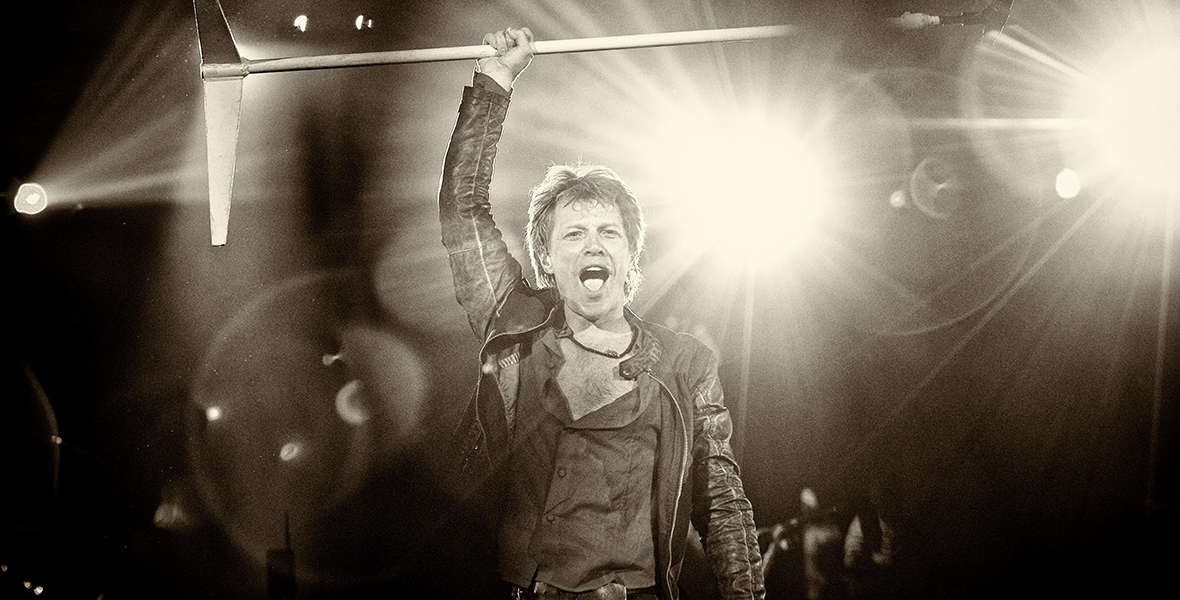 In a black and white photo, singer Bon Jovi stands onstage, raising a microphone on a stand above his head with his right hand. He is clad in a leather jacket, a partially unbuttoned shirt, and trousers. Three headlights behind him cast beams.