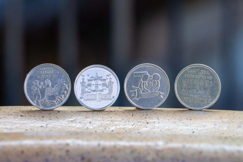 Four commemorative Disney coins displayed in a row on a stone surface. The coins feature designs from 'Tiana's Bayou Adventure,' 'Haunted Mansion,' Disneyland's 70th anniversary, and a welcome message.