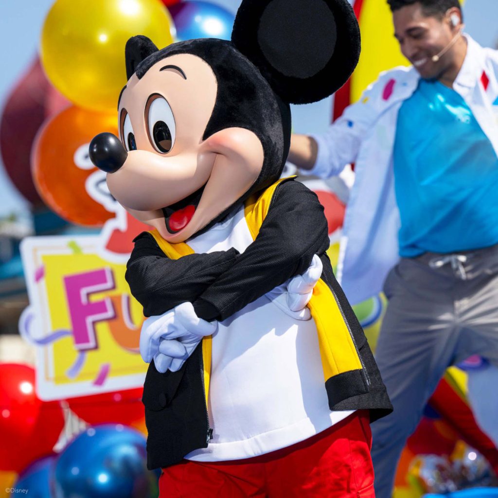 Mickey Mouse is joyfully crossing his arms while dancing in front of colorful balloons and a playful background, with a smiling performer in the background. 