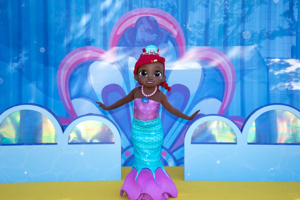 The image features a colorful statue of a young mermaid character set against a bright and vibrant backdrop. She is wearing a pink and purple top with a blue mermaid tail adorned with scales, transitioning into pink fins. Her hair is styled with a red headscarf, and she is accessorized with a pink pearl necklace and a blue seashell pendant. The background is decorated with aquatic-themed graphics, including blue and pink seashell patterns and bubble-like designs, giving the scene an underwater fantasy atmosphere. The mermaid's pose is open and welcoming, as if greeting visitors.