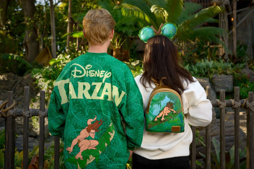 The image shows two people standing with their backs to the camera, showcasing their Disney Tarzan-themed apparel. The person on the left is wearing a green jacket with a leafy jungle pattern and the text "Disney Tarzan" in large yellow letters. Below the text, there is an illustration of Tarzan in a dynamic pose, swinging through the jungle. The person on the right is wearing a white top and a small backpack featuring a similar Tarzan illustration. They are also wearing green Mickey Mouse ears with a jungle theme. The background suggests a tropical or jungle setting, with lush greenery and a wooden fence, enhancing the overall theme. 