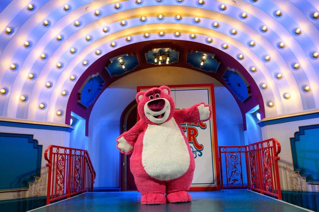 The image features Lotso from Disney's "Toy Story 3," standing on a stage. The setting is vibrant and colorful, with an arched backdrop adorned with multiple light bulbs, creating a theatrical or carnival-like atmosphere. Lotso is in the center of the stage, with his arms open wide, appearing cheerful and welcoming. 
