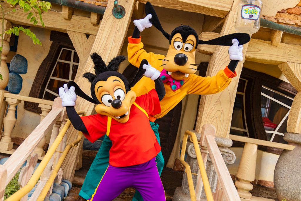 The image features Max and Goofy, two beloved Disney characters, posing playfully in front of a whimsical, cartoon-like house. Max, wearing a red shirt and purple pants, is in the foreground with an energetic and enthusiastic pose. Behind him, Goofy, dressed in his classic orange shirt and green pants with a pink and teal bowtie, is mimicking Max's pose, adding to the playful atmosphere. 