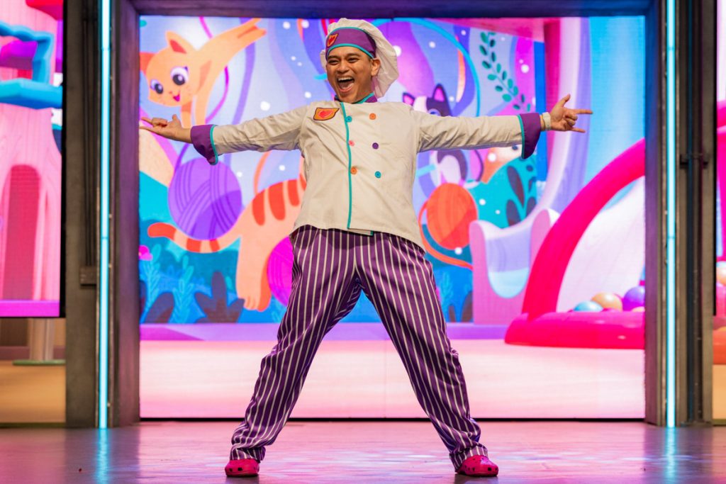 The image shows a performer on stage dressed in a colorful chef outfit. The performer is wearing a white chef's jacket with colorful buttons and accents, paired with purple and white striped pants and bright pink shoes. The chef's hat and headband match the outfit, adding to the playful appearance. The performer is standing with their arms outstretched in an energetic and welcoming pose, smiling broadly. 