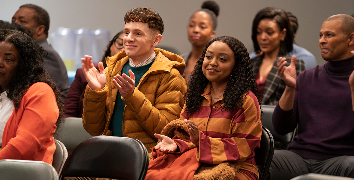In a scene from the television show Abbott Elementary, Jacob Hill (left), played by Chris Perfetti and Janine Teagues (right), played by Quinta Brunson, are sitting in chairs within an audience space, clapping with pride. Perfetti wears a checkered shirt, a green sweater, and a mustard puffer jacket, while Brunson wears an orange and red striped dress. Other audience members are also seen joining the applause.