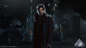 Cassian Andor stands facing the camera, standing in front of crates, barrels, and other supplies. He is mostly in darkness except for a light that shines across his face.