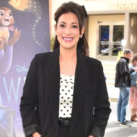 Angelique Cabral smiles at the camera and stands outside on the grass in front of a Disney Animation Studios Wish movie poster on the Walt Disney Studios lot. The theater and other guests are visible in the background behind her.