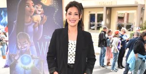 Angelique Cabral smiles at the camera and stands outside on the grass in front of a Disney Animation Studios Wish movie poster on the Walt Disney Studios lot. The theater and other guests are visible in the background behind her.