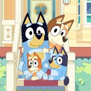 In a scene from the series Bluey, Chilli, Bandit, Bluey, and Bingo, a family of Australian Cattle dogs, are sitting on the porch of their home. Chilli, the family’s mother, and Bingo, the family’s youngest, are Red Heelers, and Bandit, the family’s father, and Bluey, the family’s first child, are Blue Heelers. All members are looking calmly at the camera, but Bingo is frightened.