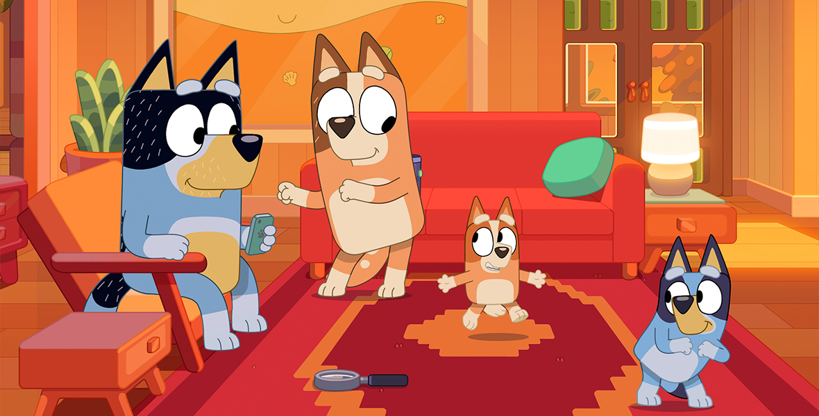 In a scene from the series Bluey, Chilli, Bandit, Bluey, and Bingo, a family of Australian Cattle dogs, are playfully dancing in their living room. Chilli, the family’s mother, and Bingo, the family’s youngest, are Red Heelers, and Bandit, the family’s father, and Bluey, the family’s first child, are Blue Heelers. The living room has a red and orange carpet, orange chairs, and orange-tinted walls.