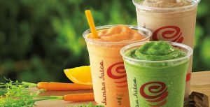 A colorful image featuring three Jamba Juice smoothies in clear plastic cups with the Jamba Juice logo. The smoothies are in various flavors: a bright orange one with a yellow straw, a green one, and a light brown one. Fresh carrots, an orange slice, and pieces of chocolate are placed around the smoothies, against a blurred green background.