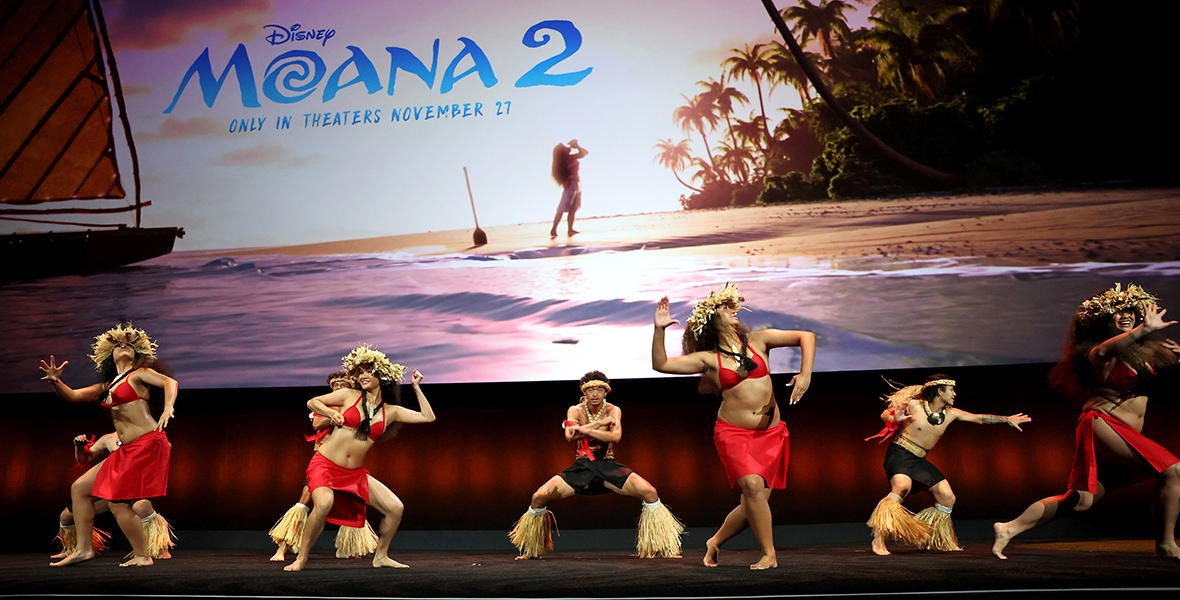 Polynesian dancers perform onstage during the Walt Disney Studios Presentation at Cinemacon. Behind them is a teaser image for Moana 2, featuring the film’s title superimposed over the image of a beach, where Moana stands on the sand, blowing into a conch shell.