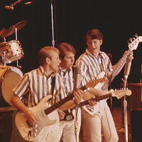 In a scene from the upcoming documentary The Beach Boys, the band is performing onstage circa 1964 in California. Dennis Wilson is seen playing the drums at the back, positioned higher than the rest of the band. At center stage are Al Jardine, Carl Wilson, Brian Wilson, and Mike Love. They are all singing, and (with the exception of Love) playing guitars. The band members are dressed in white and grey striped short-sleeve shirts and white pants.
