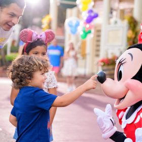 A father and two kids, a boy in a blue shirt and a girl wearing pink Minnie ears, are visiting with Minnie Mouse. Minnie is kneeling, and the boy is reaching out towards her nose.