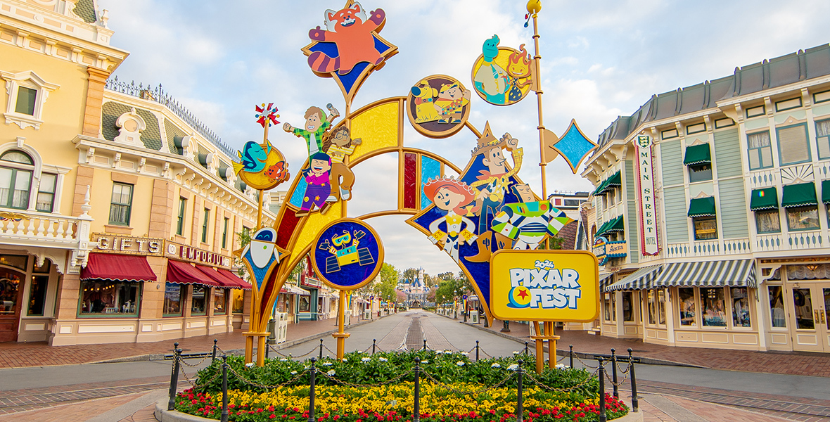 Colorful Pixar-inspired sculpture adorns flowered roundabout at Disneyland's Main Street, U.S.A. From left to right, the gold sculpture features the robot EVE from WALL-E; Dory and Nemo from Finding Nemo; Miriam, Abby, Priya, and Red Panda Mei from Turning Red; the robot WALL-E from WALL-E; Russell and Doug from Up; Wade and Ember from Elemental; and Woody, Jessie, and Buzz Lightyear from Toy Story. In front of the sculpture, a sign reads “2024 Pixar Fest.” In the background are shops and restaurants, with the Sleeping Beauty Castle in the distance.