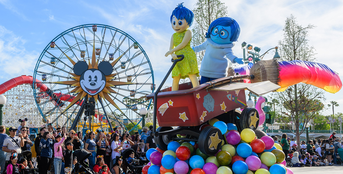 In the Better Together: A Pixar Pals Celebration parade, characters Joy and Sadness from Inside Out are in a life-size Rocket Wagon, which stands on Memory Orbs. Crowds of excited spectators line the parade route, capturing the magic with cameras. Further in the background stands the iconic Pixar-Pal-A-Round attraction. Behind that is the Incredicoaster, featuring red enclosed tunnels.