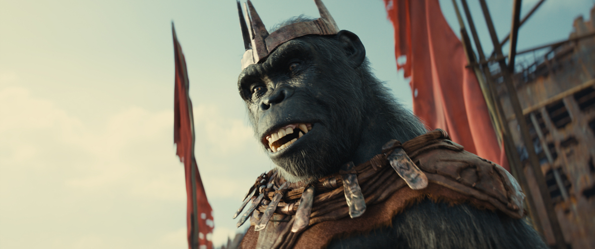 In a scene from Kingdom of the Planet of the Apes, Proximus Caesar (Kevin Durand), an ape leader, dons a bronze crown and padded armor extending from his neck to his shoulders. In the background, part of his shipwreck kingdom is visible, along with two red flags.