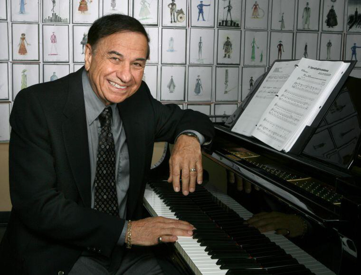 Richard M. Sherman poses at the piano. Behind him are costume sketches.