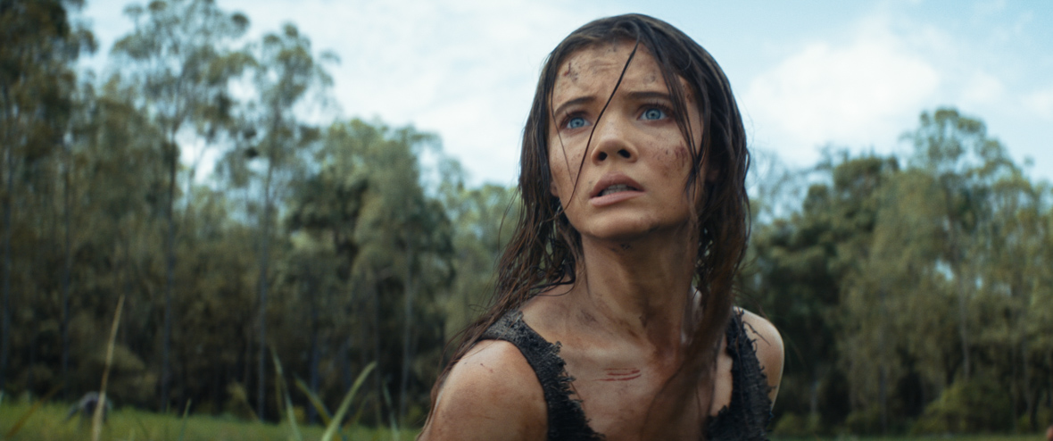 In a scene from Kingdom of the Planet of the Apes, Mae (Freya Allan) is wearing a rugged gray tank top. Looking fearful, she has scratches with blood across her face and collarbone area. In the background of the frame is a green field and the trees of a nearby forest. 
